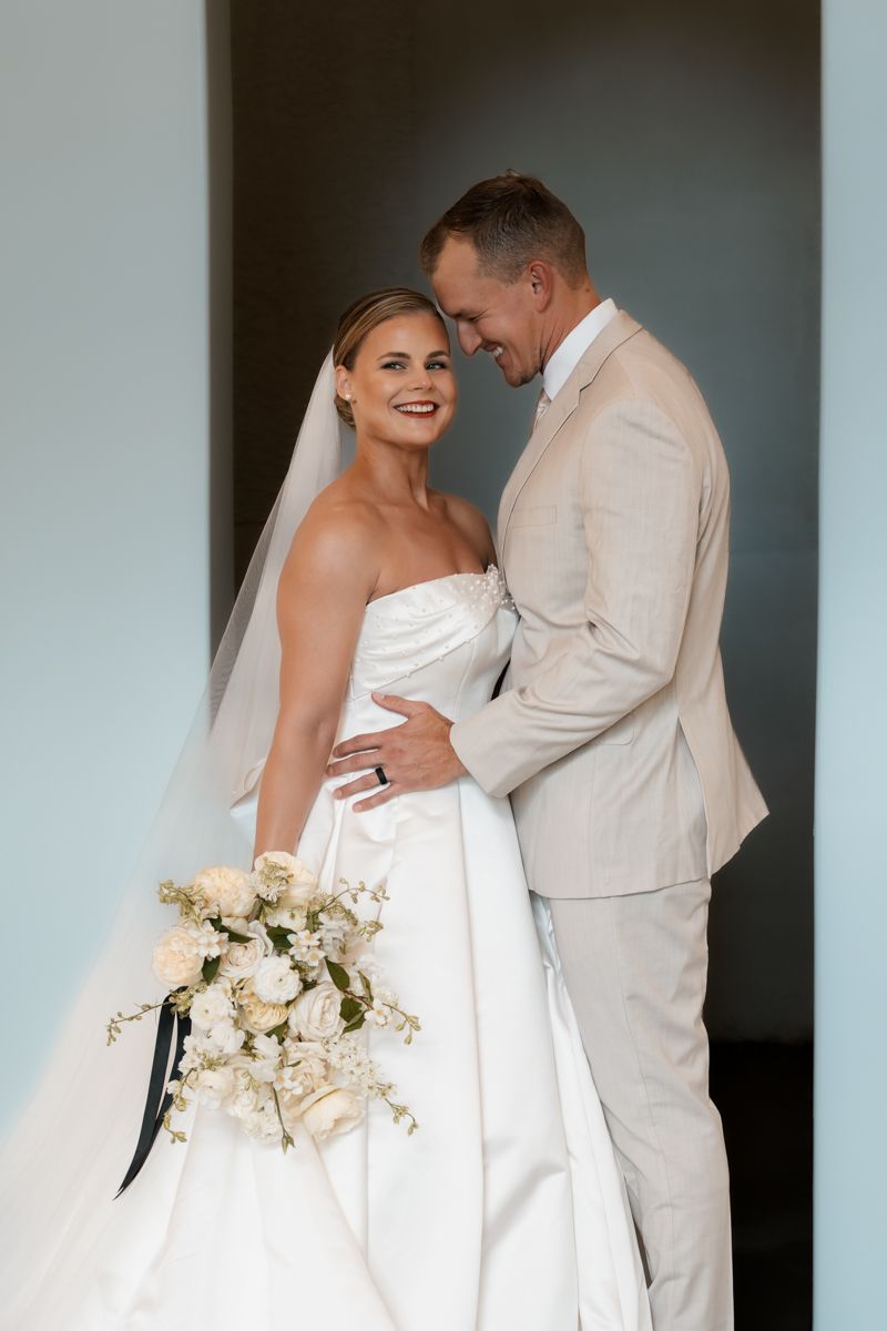 Bride and groom in light colored attire share loving embrace while holding wedding flowers.