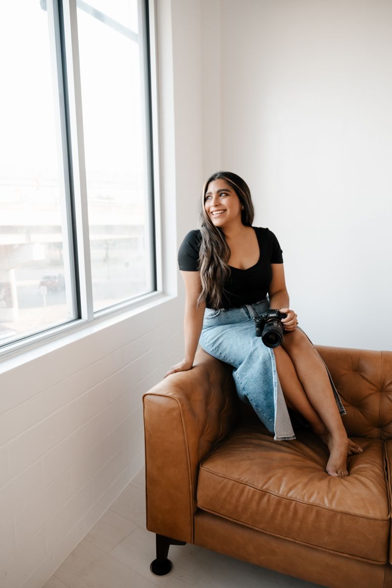 Woman in black top and jeans sits casually on brown leather couch near window in modern room.