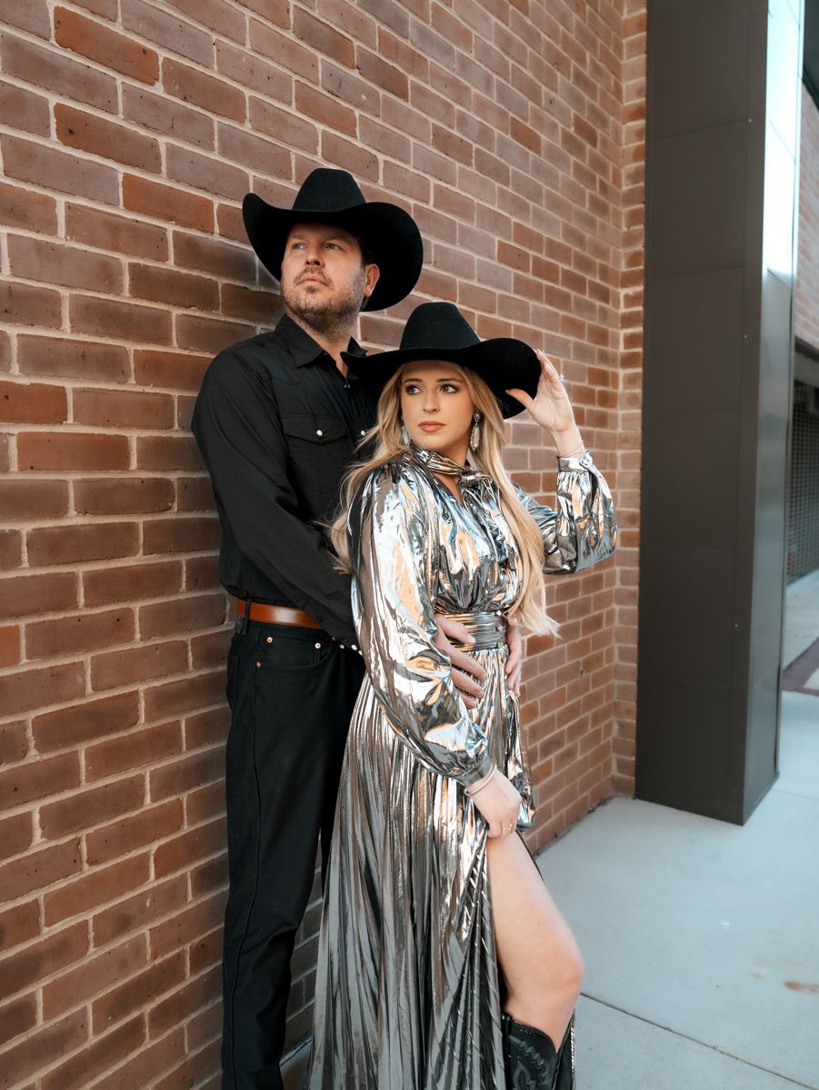 Couple in western attire poses dramatically against brick wall, woman wearing metallic dress.