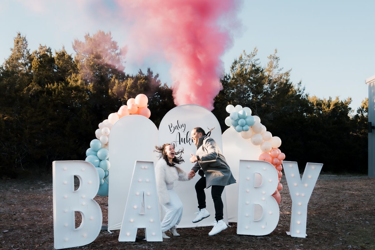 Gender reveal celebration with pink smoke and large BABY letters outdoors.