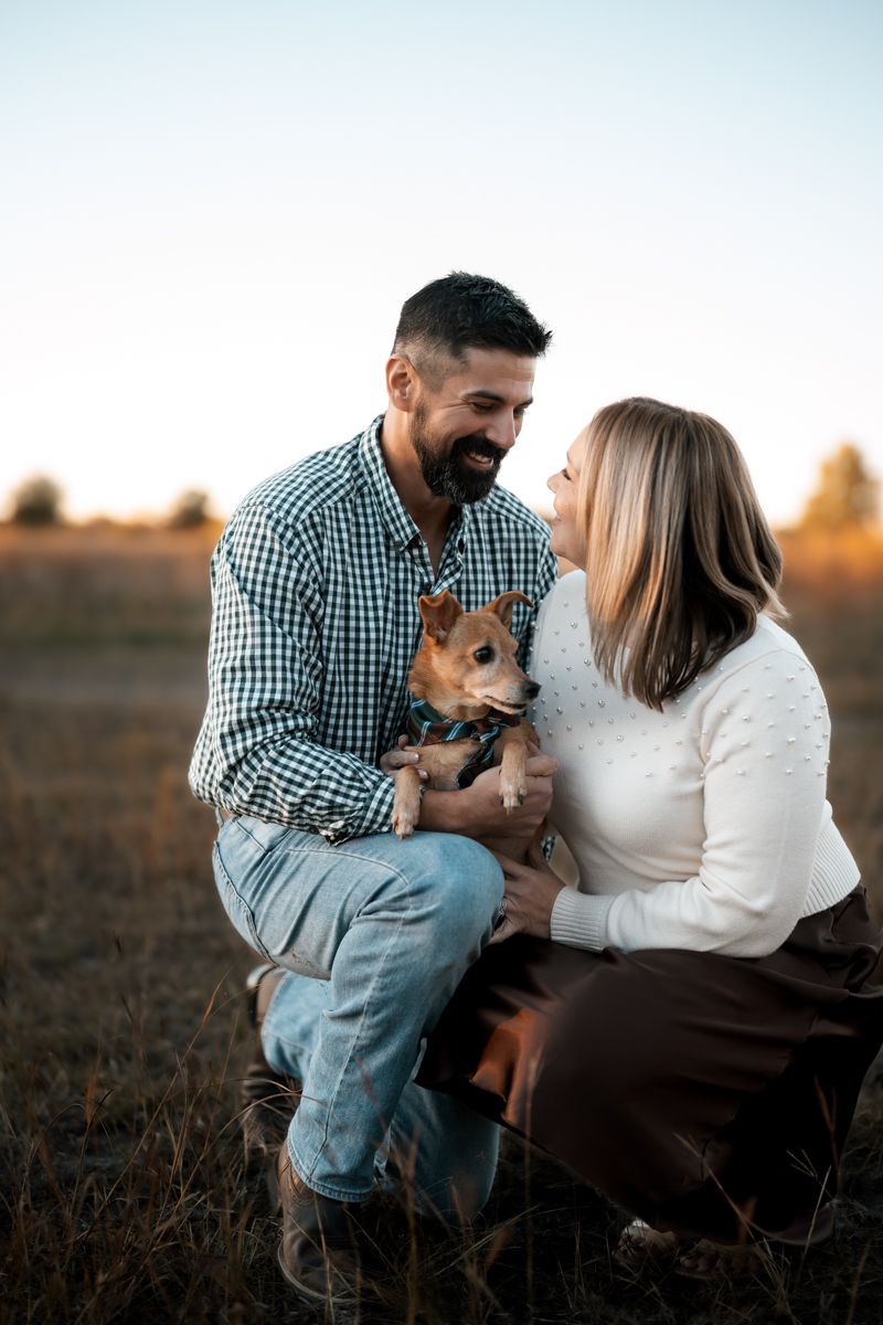 A couple shares a joyful moment with their small brown dog in a rustic outdoor setting at sunset.
