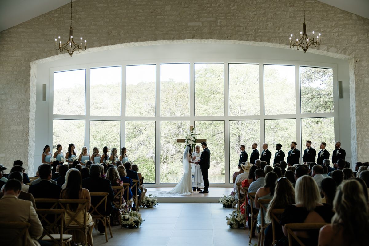 Wedding ceremony in bright white chapel with large arched windows and full audience.