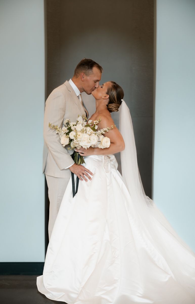 Elegant wedding portrait of couple embracing against neutral wall with white floral bouquet.