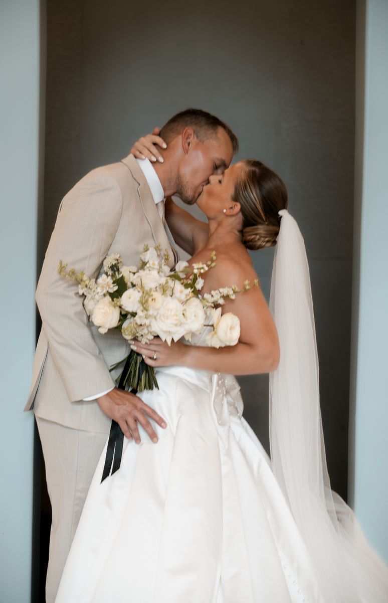 Romantic wedding couple shares intimate kiss while holding white bouquet against modern backdrop.
