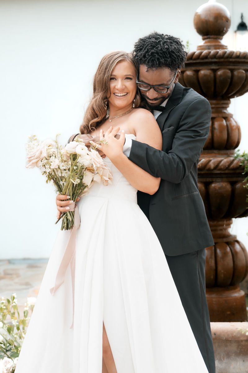 Joyful wedding couple embraces by stone fountain while holding delicate white bouquet.