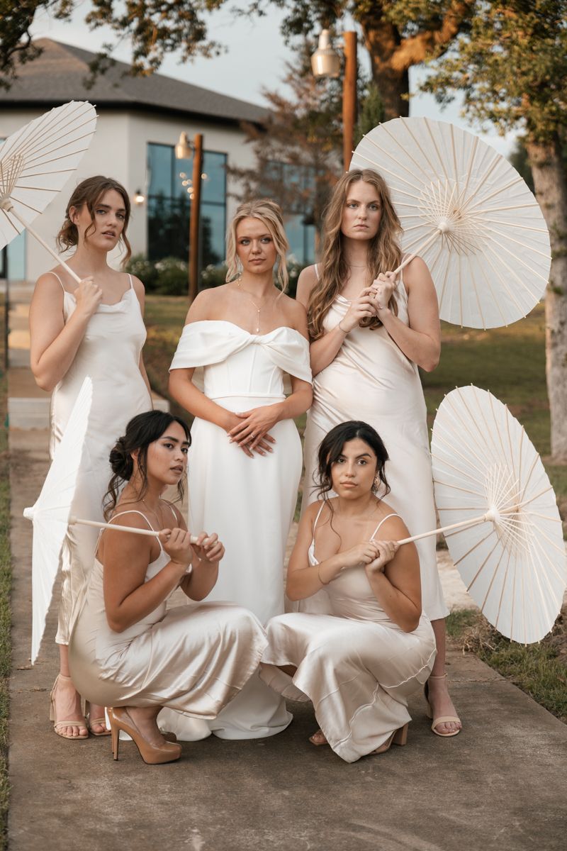 Group portrait with white parasols and coordinating cream dresses in garden setting.
