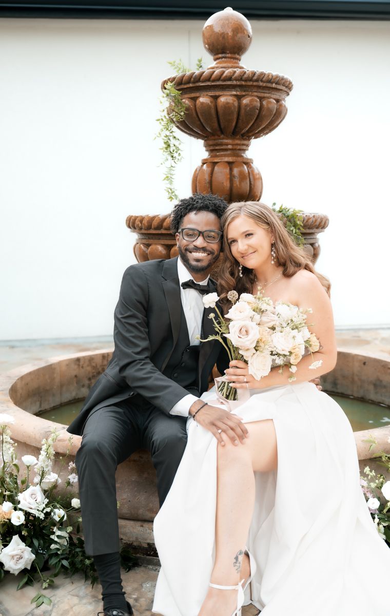 Newlyweds pose elegantly by ornate stone fountain with white floral bouquet.