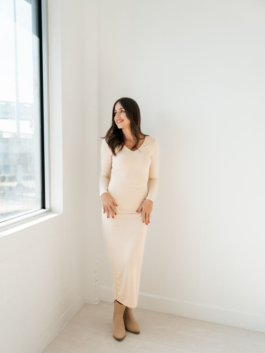Woman in cream colored long-sleeve maxi dress standing by bright window.