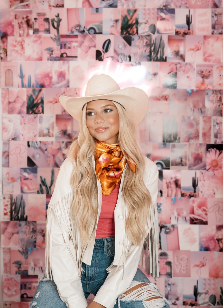 Western style portrait with cowboy hat, fringe jacket, and bandana against pink collage wall.