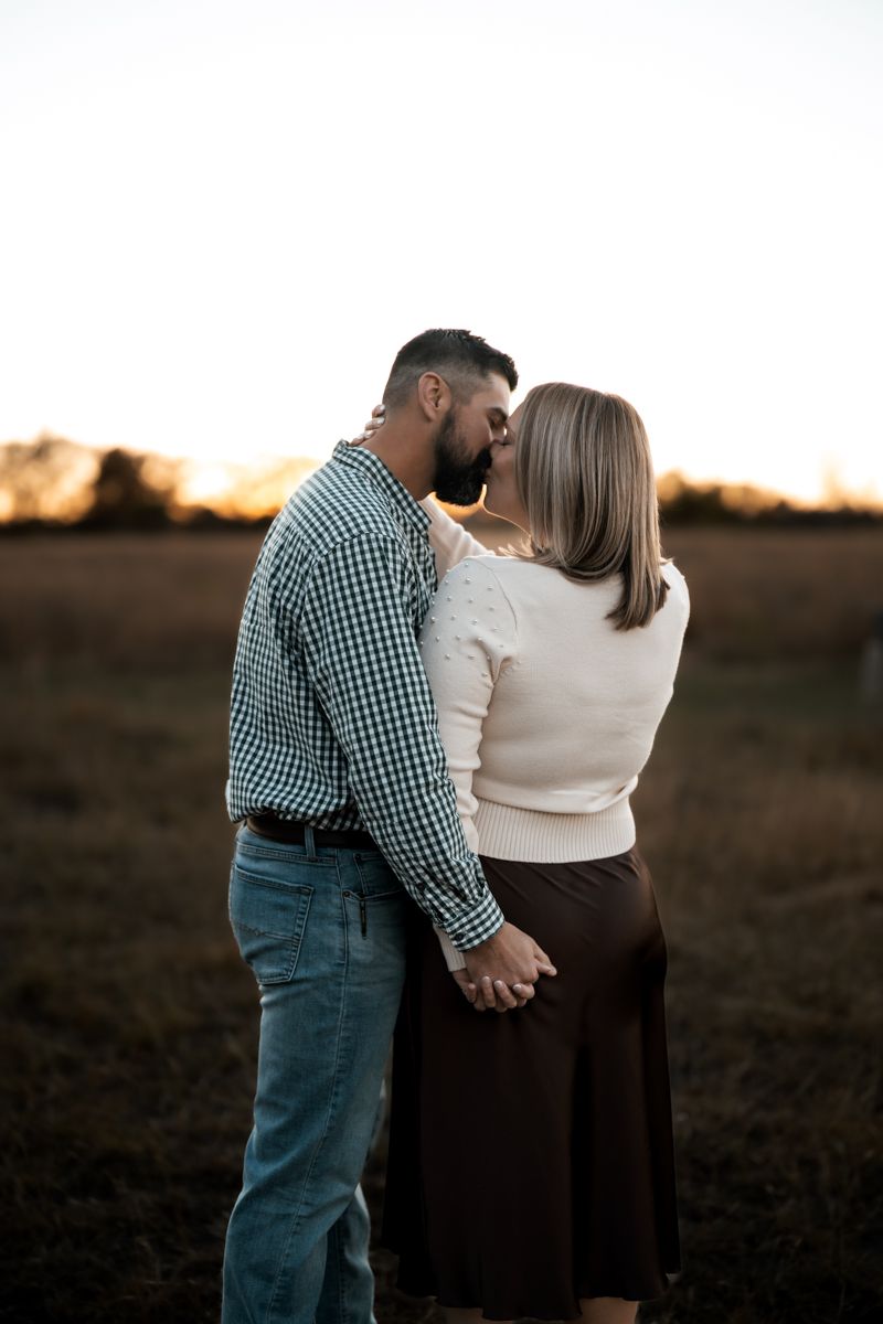 A couple shares an intimate embrace in casual attire against a sunset backdrop in an open field.