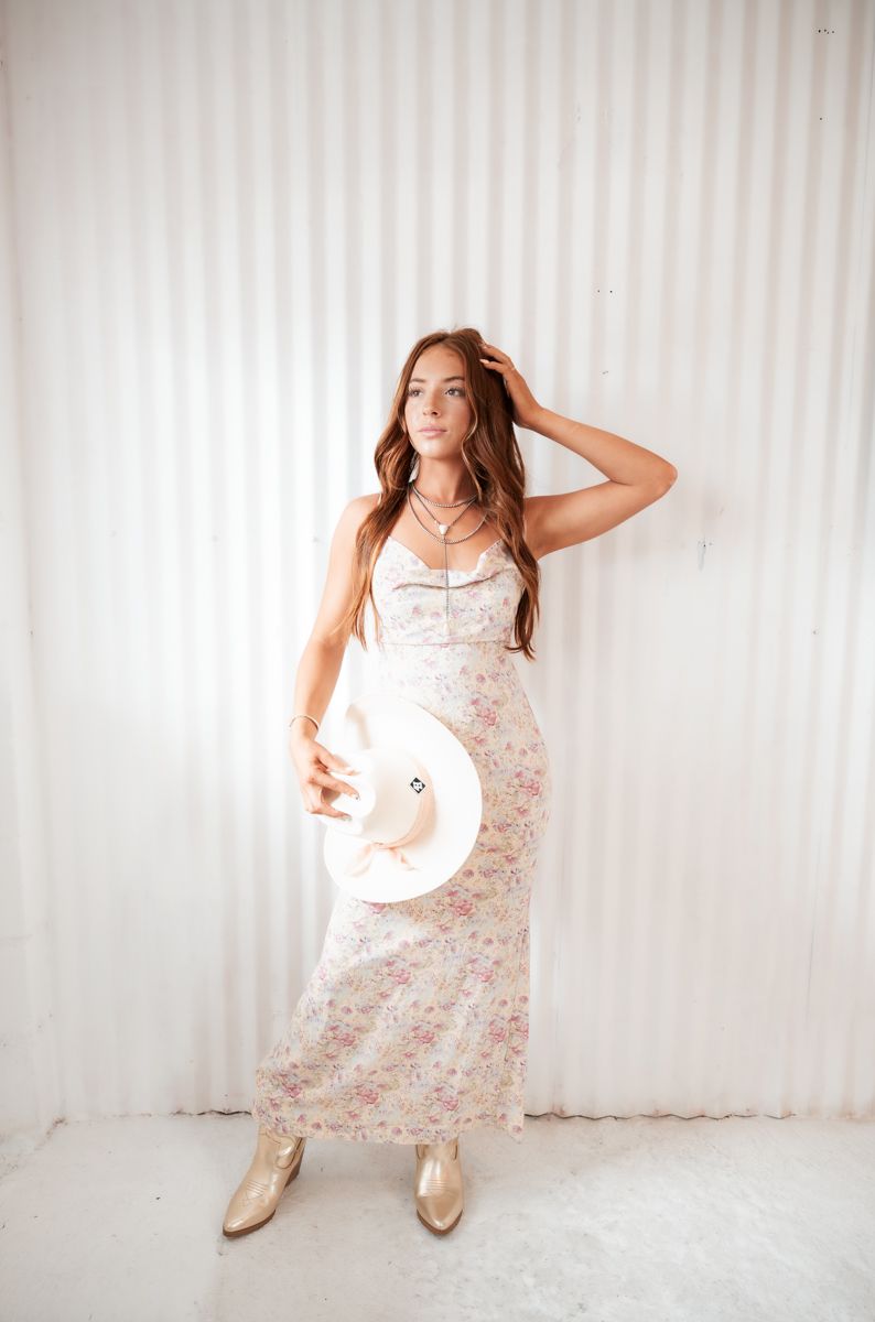 Model in floral maxi dress holds white hat while posing in front of white striped background.