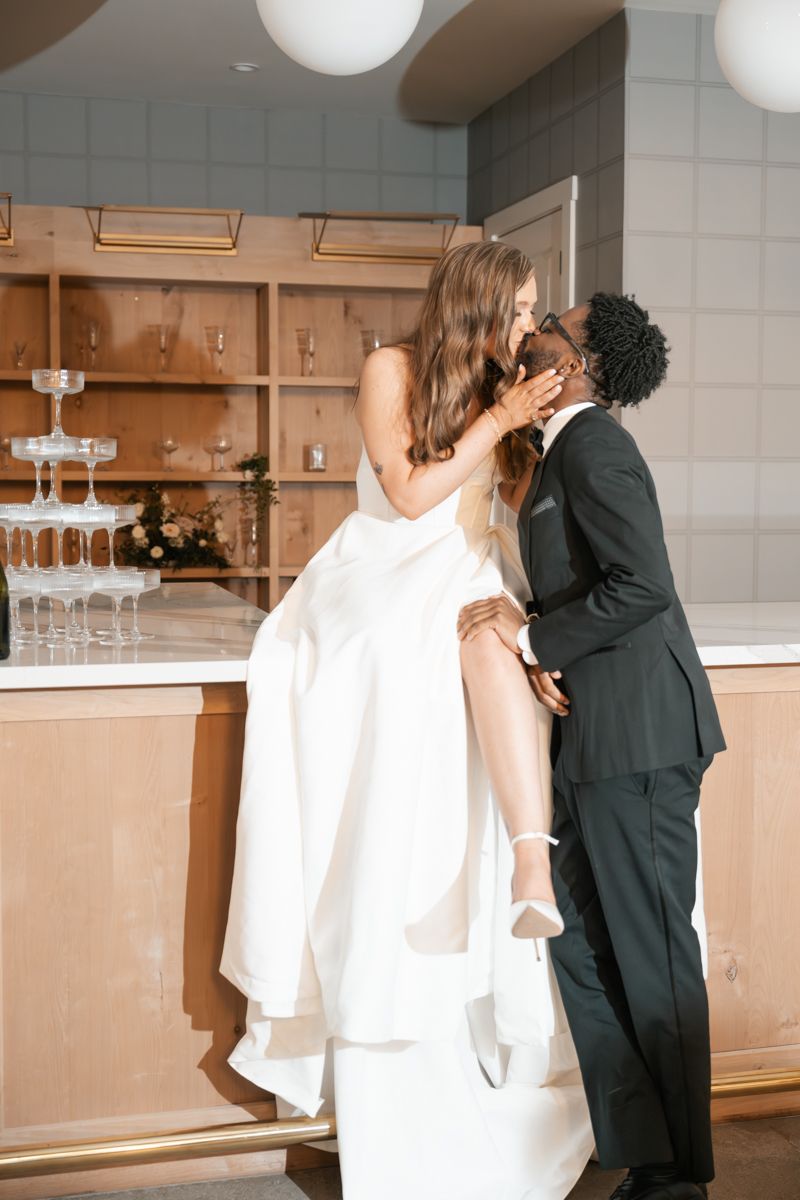 A playful wedding moment captured at a modern bar with wooden shelving and globe lights.
