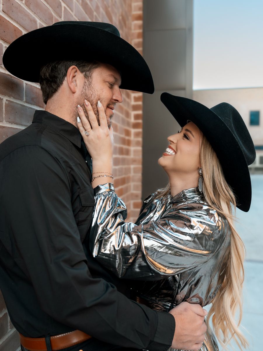 Couple in matching black cowboy hats sharing tender moment against brick wall.