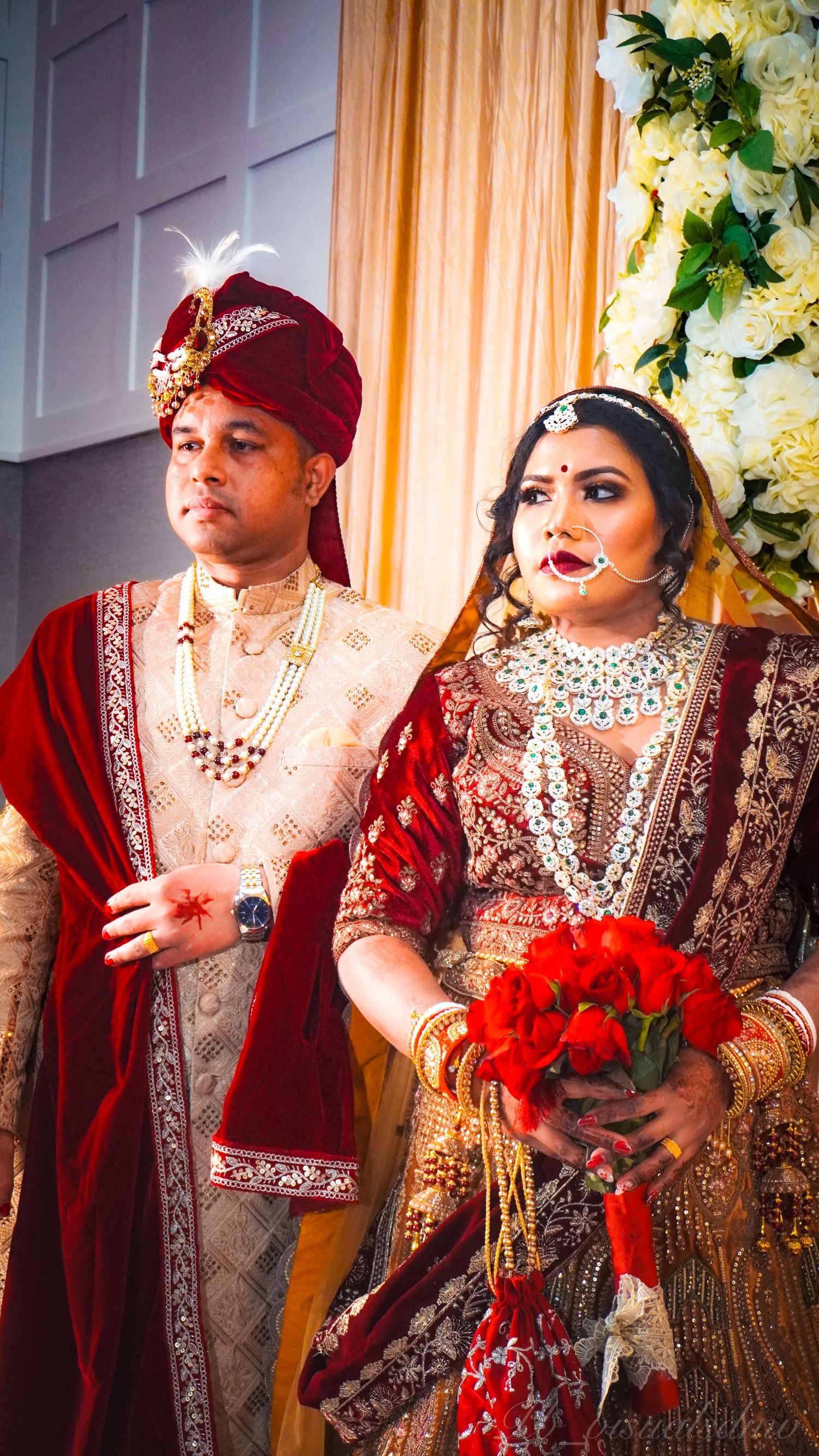 Bride and groom in traditional South Asian wedding attire standing together, bride holding bouquet of red roses against floral backdrop.
