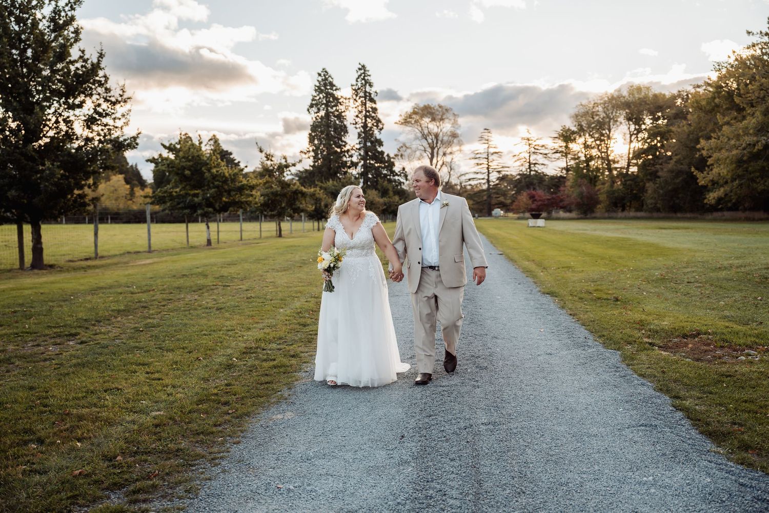 Bride and groom walk hand in hand down gravel path at sunset on wedding day.