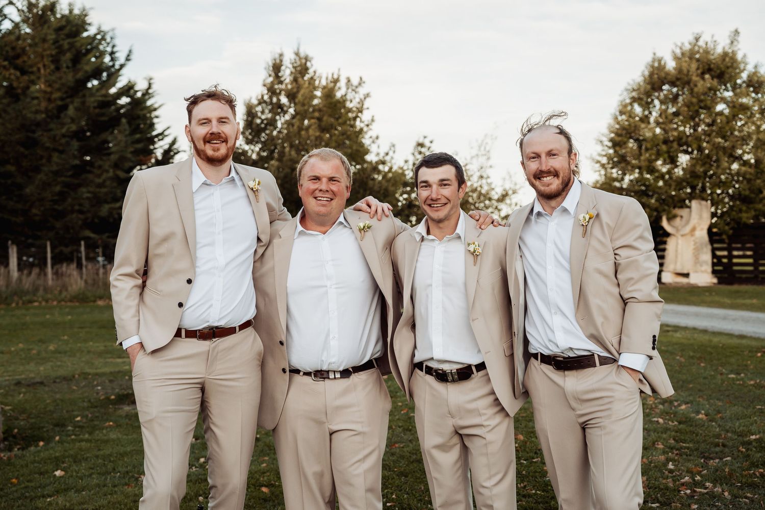Four groomsmen in matching tan suits smiling together on green lawn outdoors.