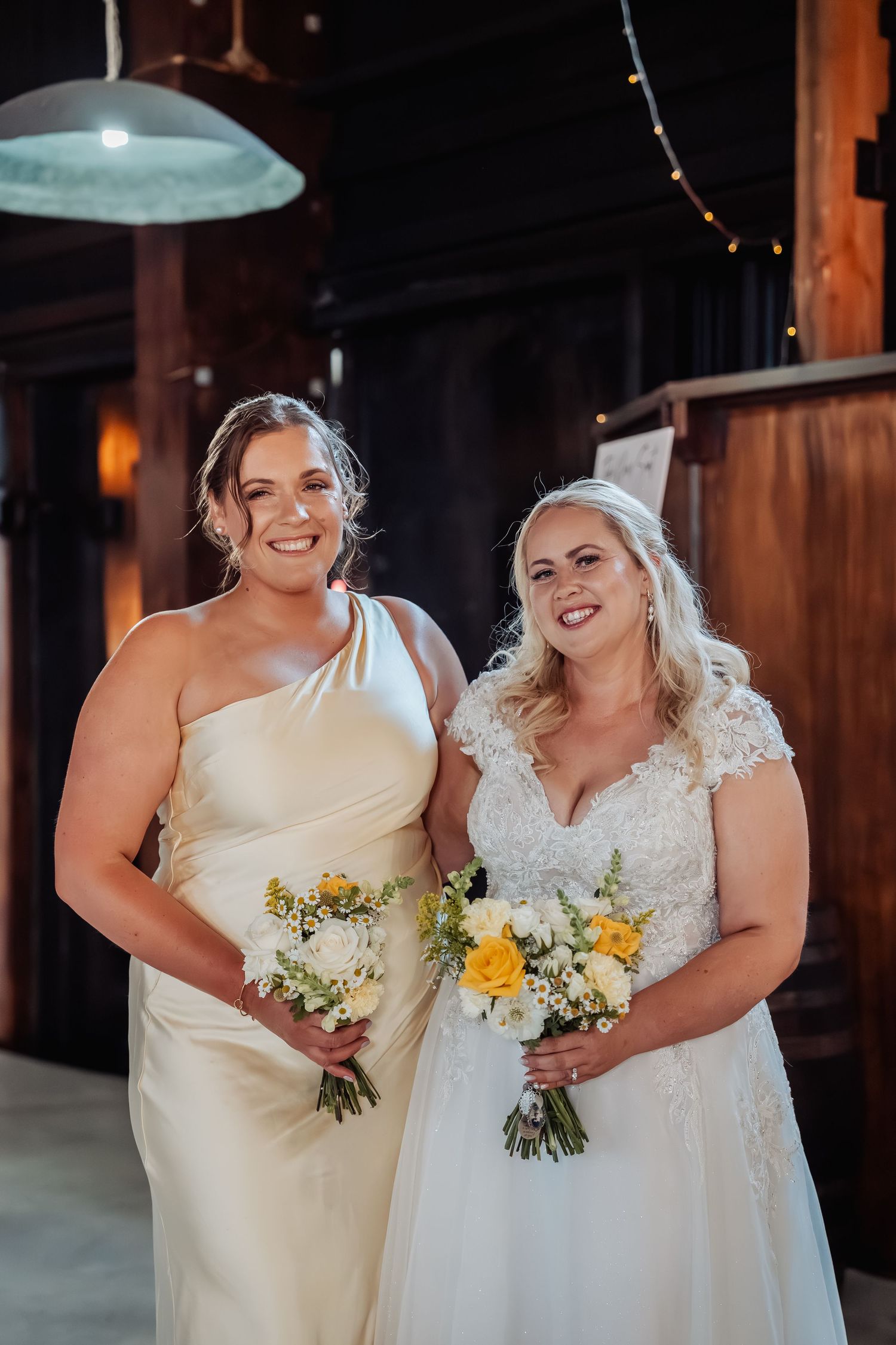 Two brides pose in a barn venue, one in a one-shoulder satin gown and one in a lace cap-sleeve wedding dress.