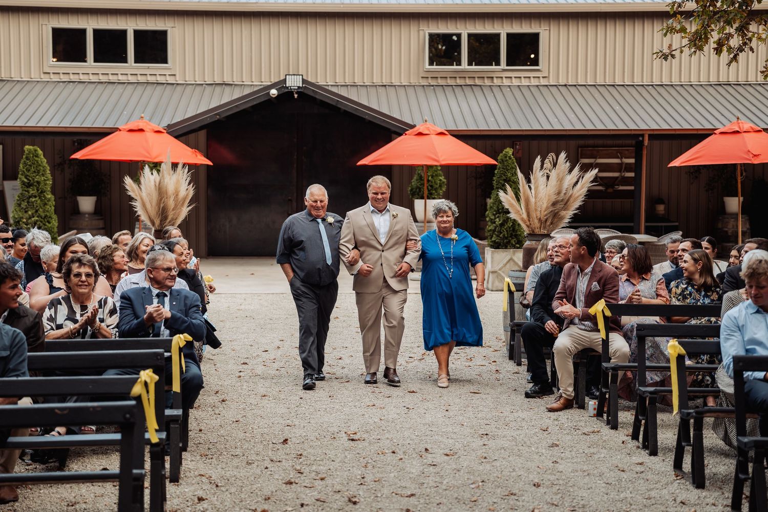 Wedding ceremony processional with elderly couple being escorted down outdoor aisle at barn venue with orange umbrellas.