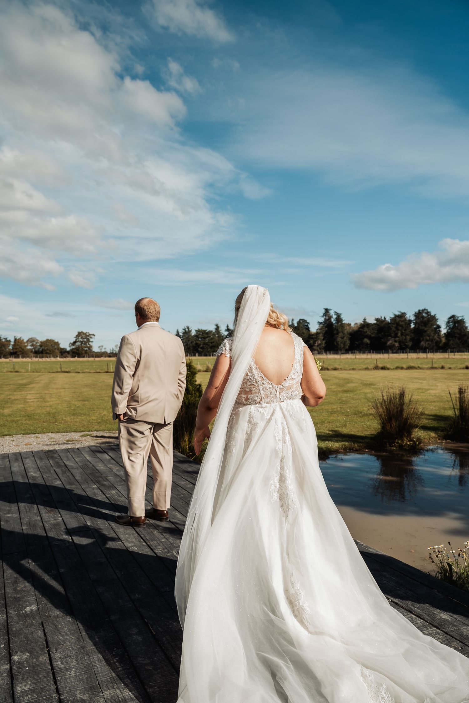 Bride and groom walking on wooden deck overlooking green fields.