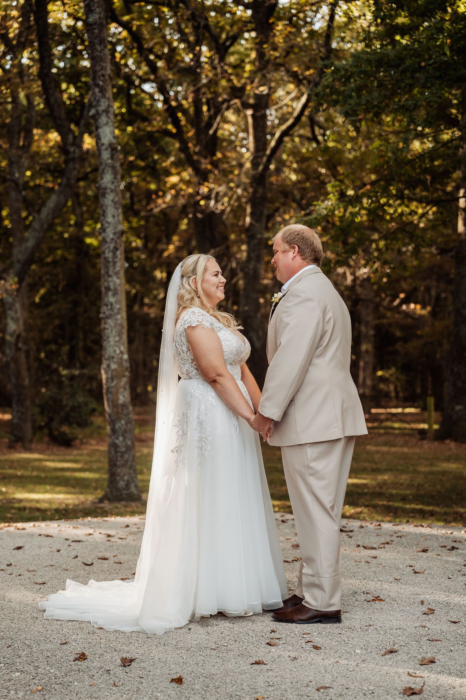 Bride in lace gown with veil and groom in tan suit hold hands and smile at each other outdoors.
