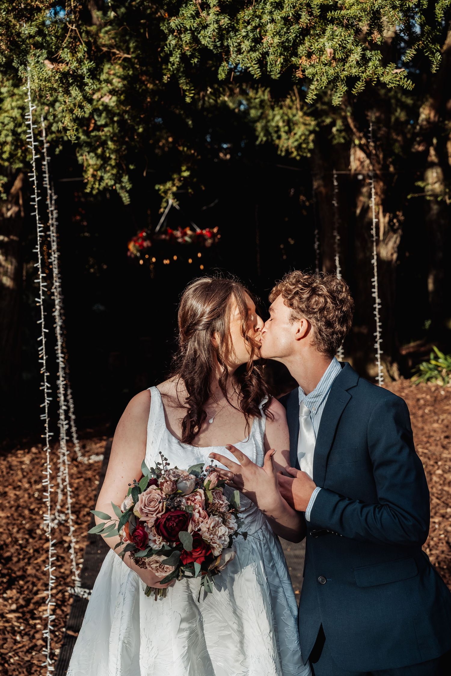 Newlyweds share first kiss surrounded by fairy lights and evergreen trees after outdoor wedding ceremony.