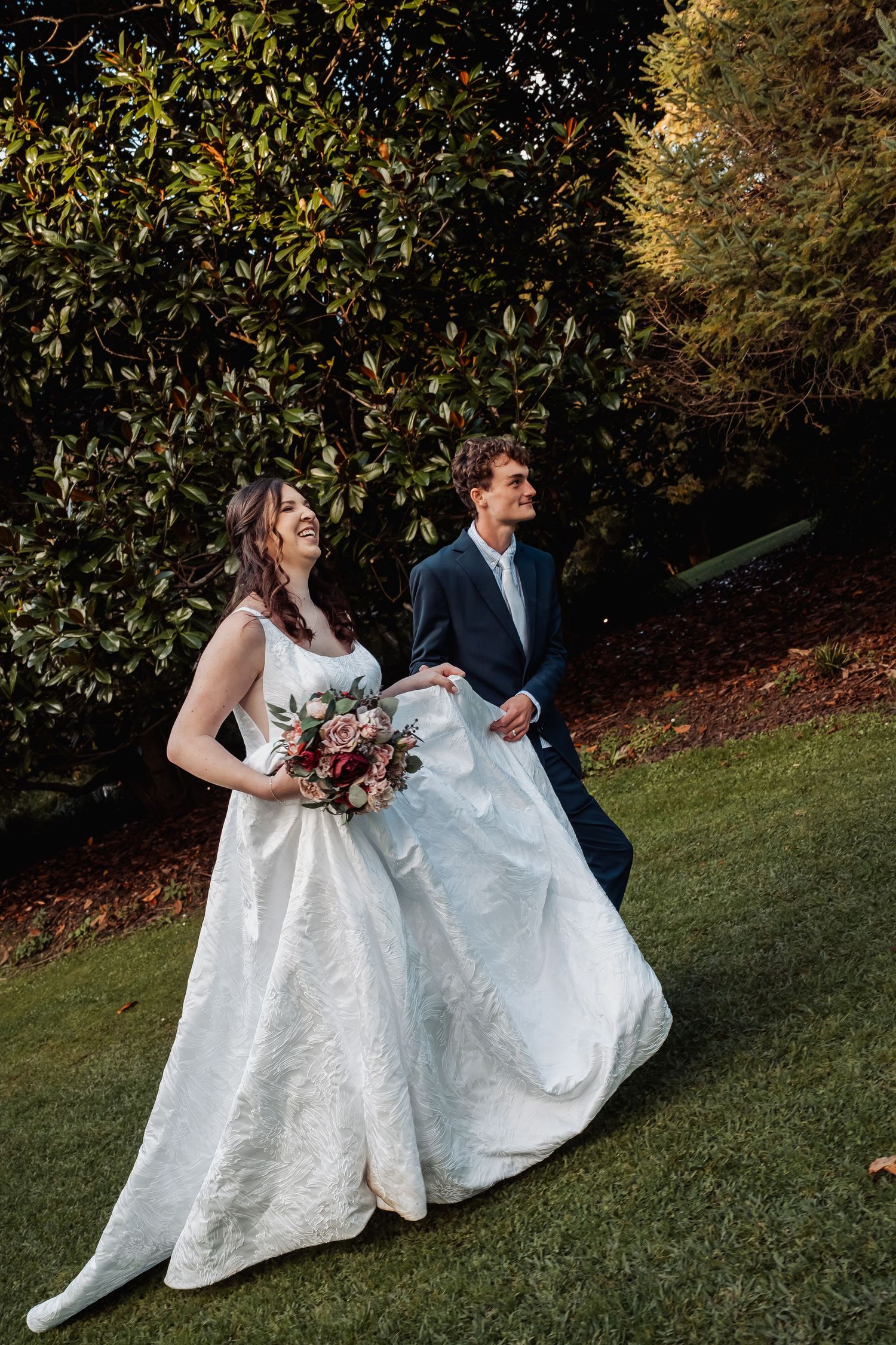 Bride and groom walking on green lawn, groom holding dress train.