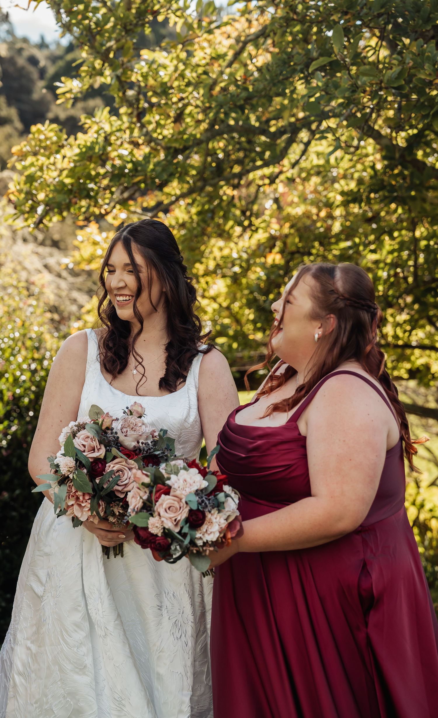 Bride in white gown laughs with bridesmaid in burgundy dress holding floral bouquets outdoors.