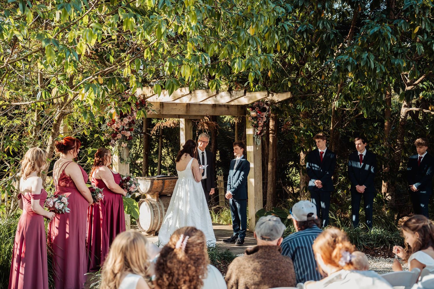 Wedding ceremony under wooden arbor with bridesmaids in burgundy and groomsmen in navy suits.