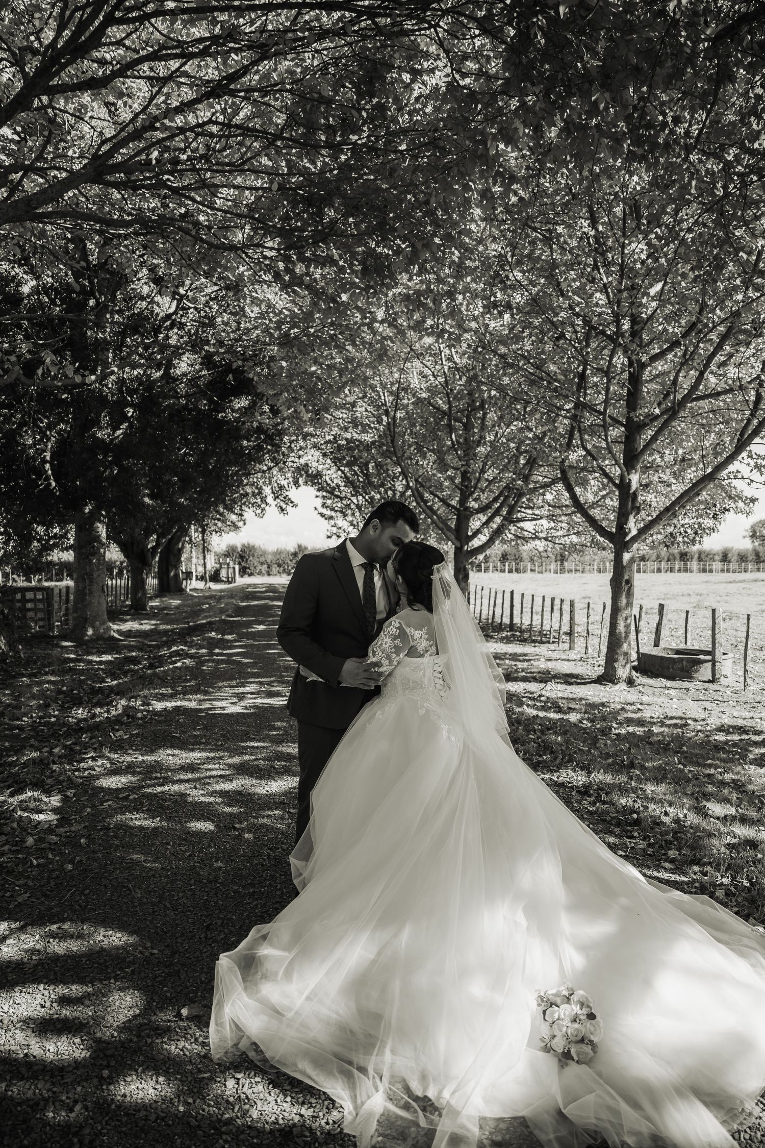 Black and white photo of bride and groom kissing along a tree-lined path.