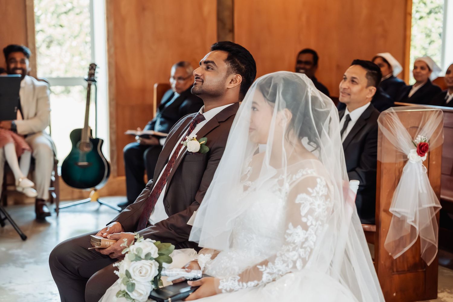 Bride and groom seated together during wedding ceremony, bride wearing lace gown and veil, guests visible in background.