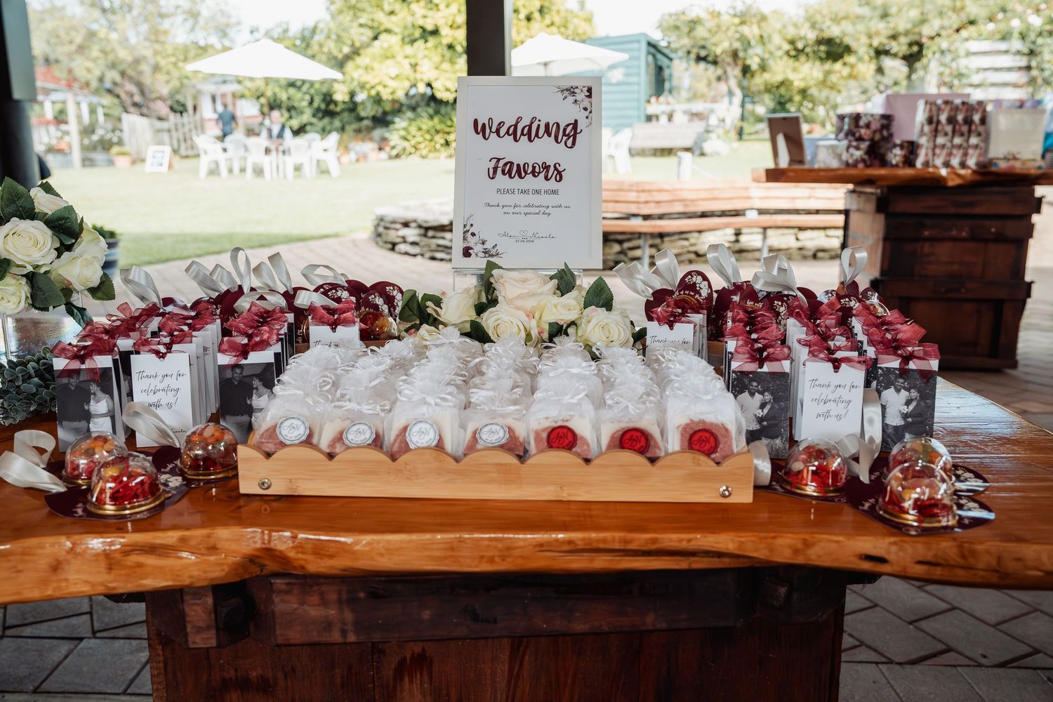 Rustic wooden table displaying elegant wedding favors with white floral arrangements and a decorative sign at an outdoor reception.