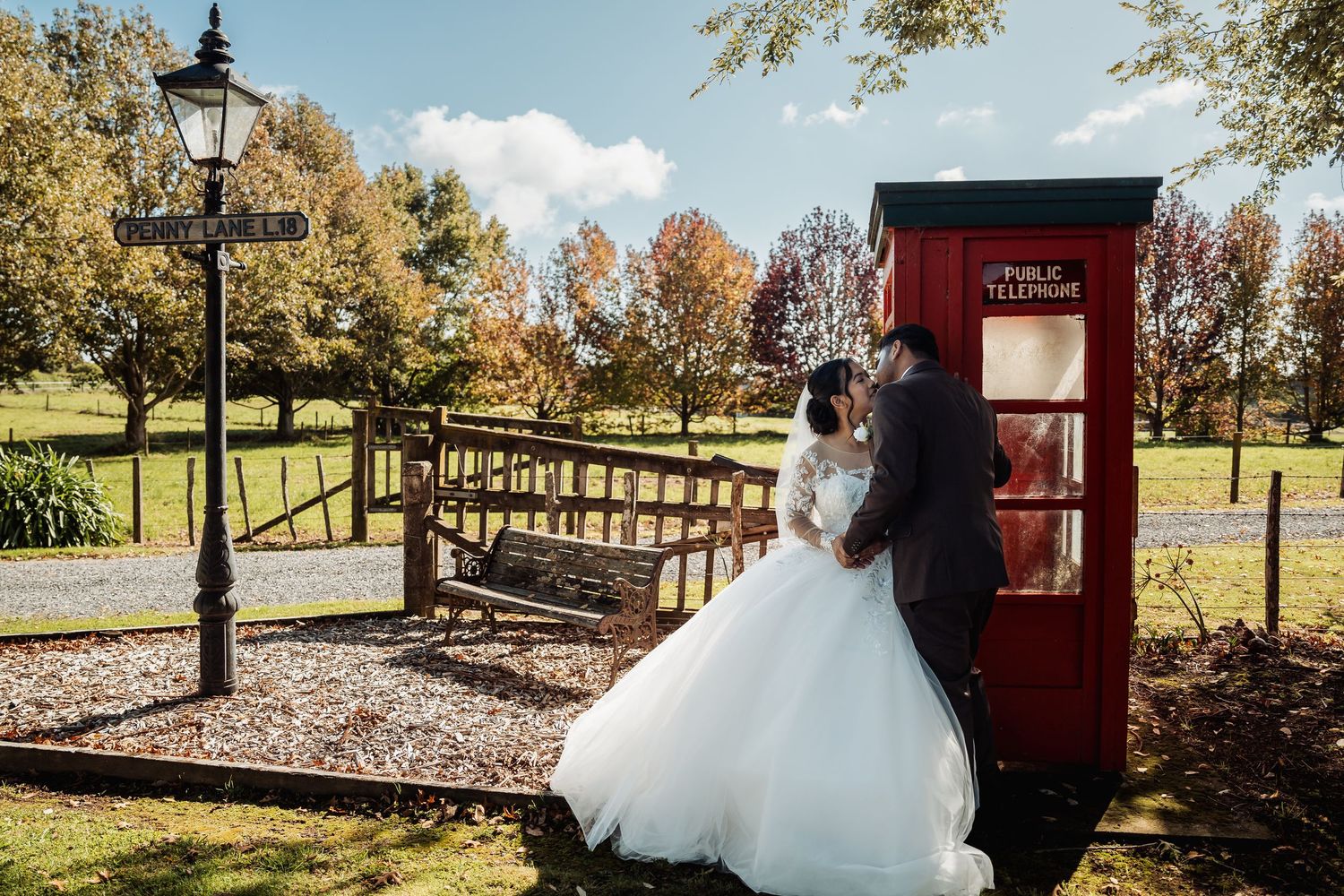 Bride and groom share a romantic kiss beside a red British phone box in an autumn park setting.
