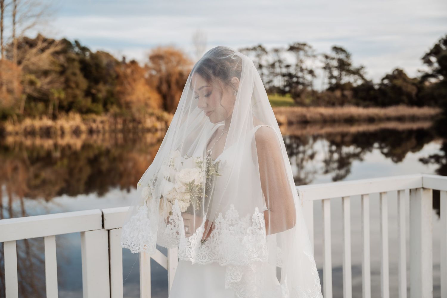 A flowing white wedding dress trails behind against a lakeside landscape with autumn trees.