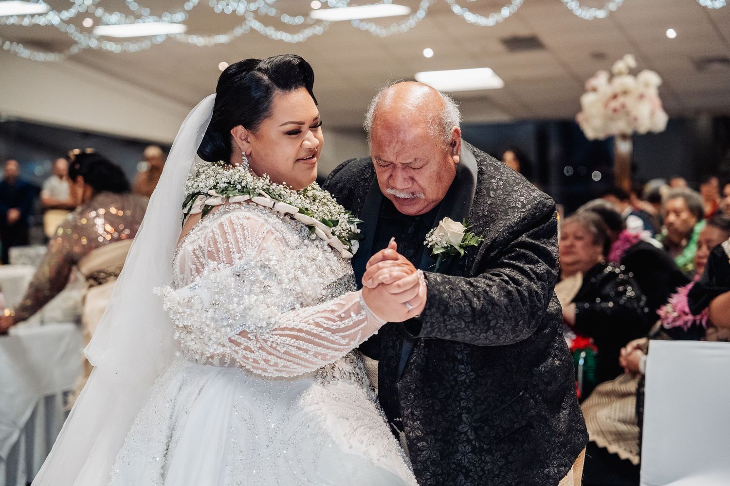 Bride in lace wedding dress dancing with elderly guest at indoor reception.