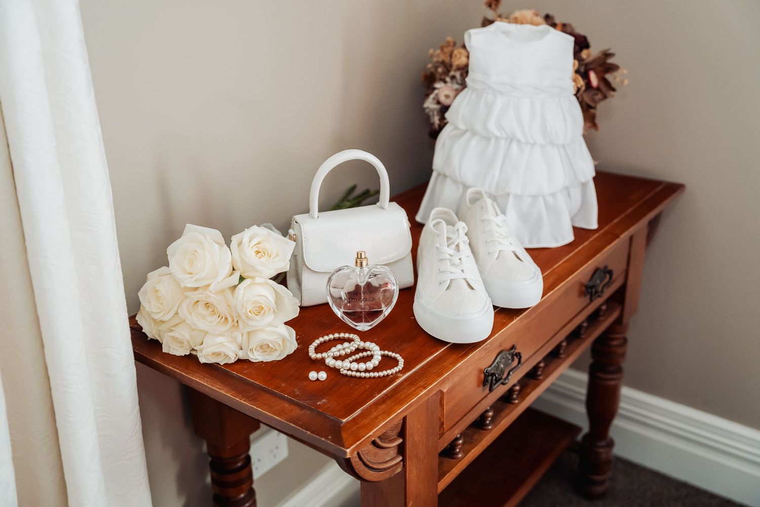 White baby dress, shoes, purse and accessories arranged on a wooden vintage table with roses for a baptism celebration.