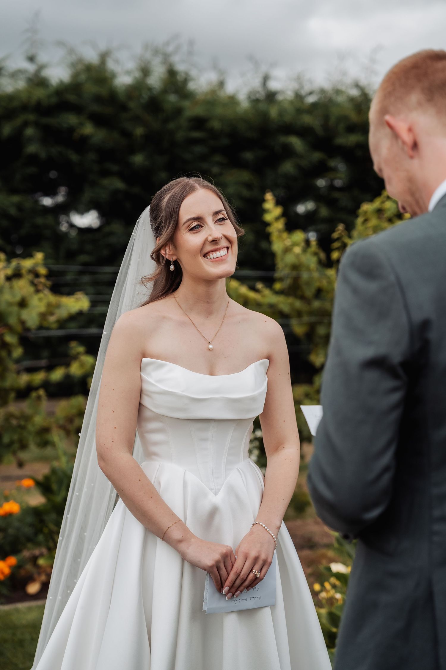 Radiant bride in a strapless white gown and veil smiling joyfully at her groom during an outdoor wedding ceremony.