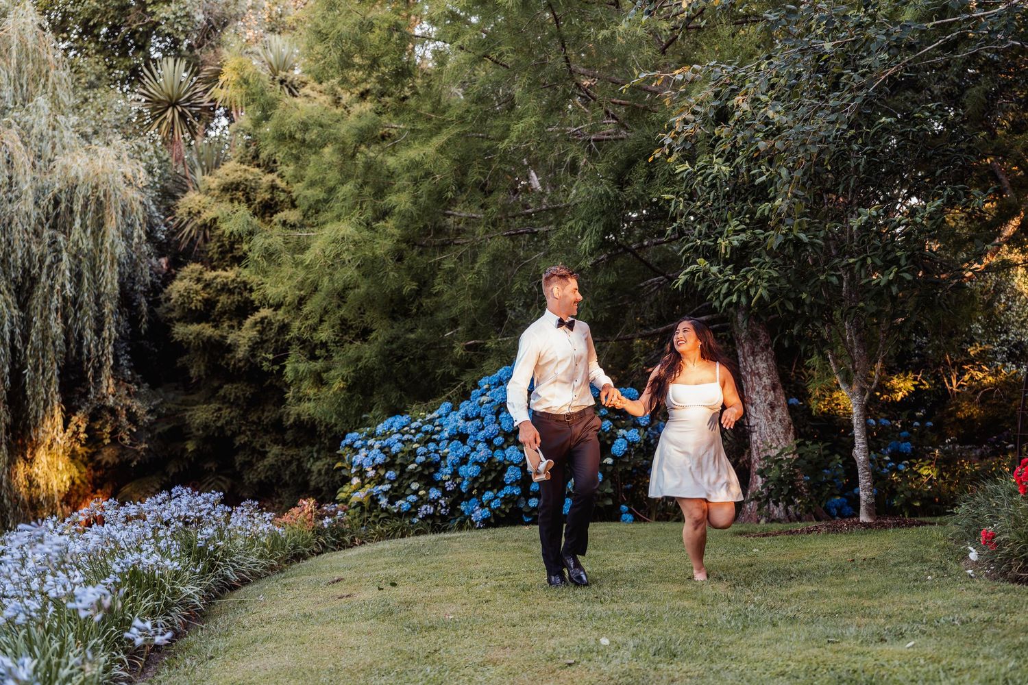 Joyful couple holding hands and walking through a colorful garden at golden hour.