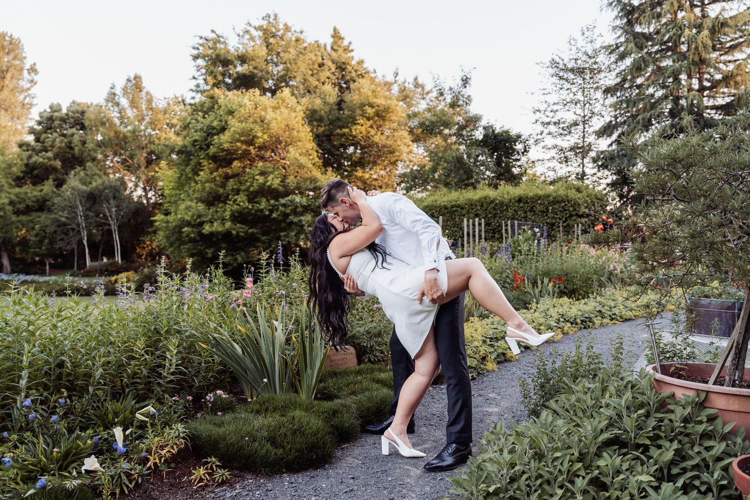 Groom joyfully lifting bride in white dress in a lush garden during wedding photo session.