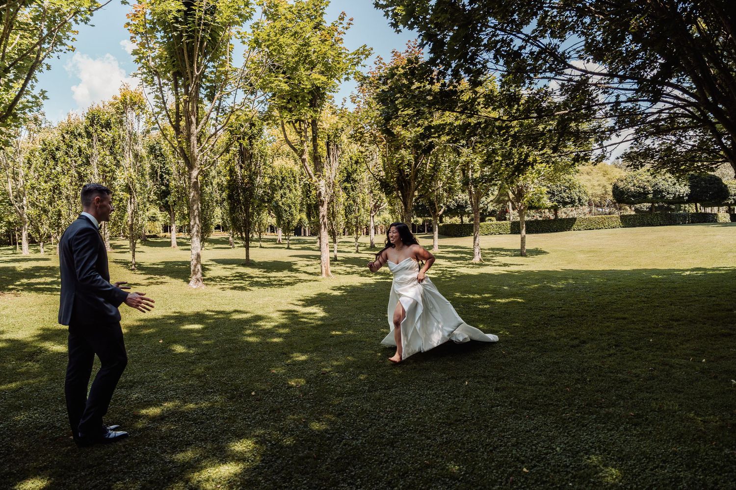 Bride in white gown running toward groom in dark suit on lush green lawn surrounded by tall trees.