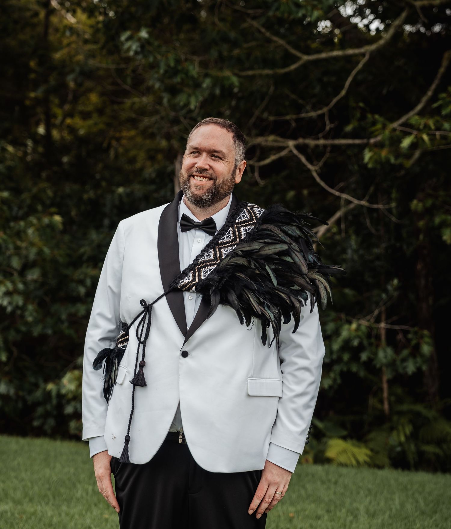 Groom in white tuxedo with black feather Maori korowai cloak smiling outdoors.