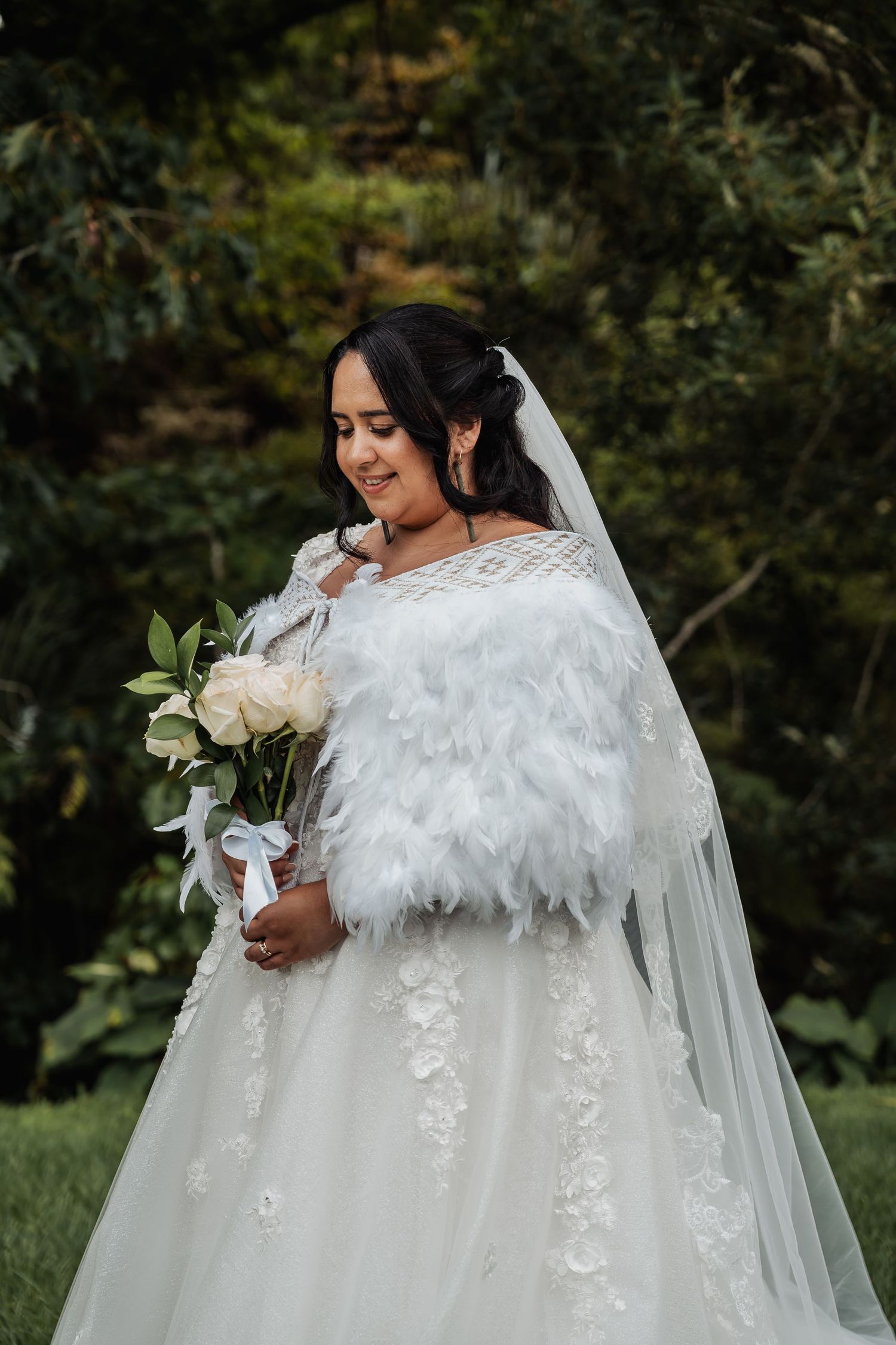 Bride in white lace gown with feather wrap holding white rose bouquet in garden setting.