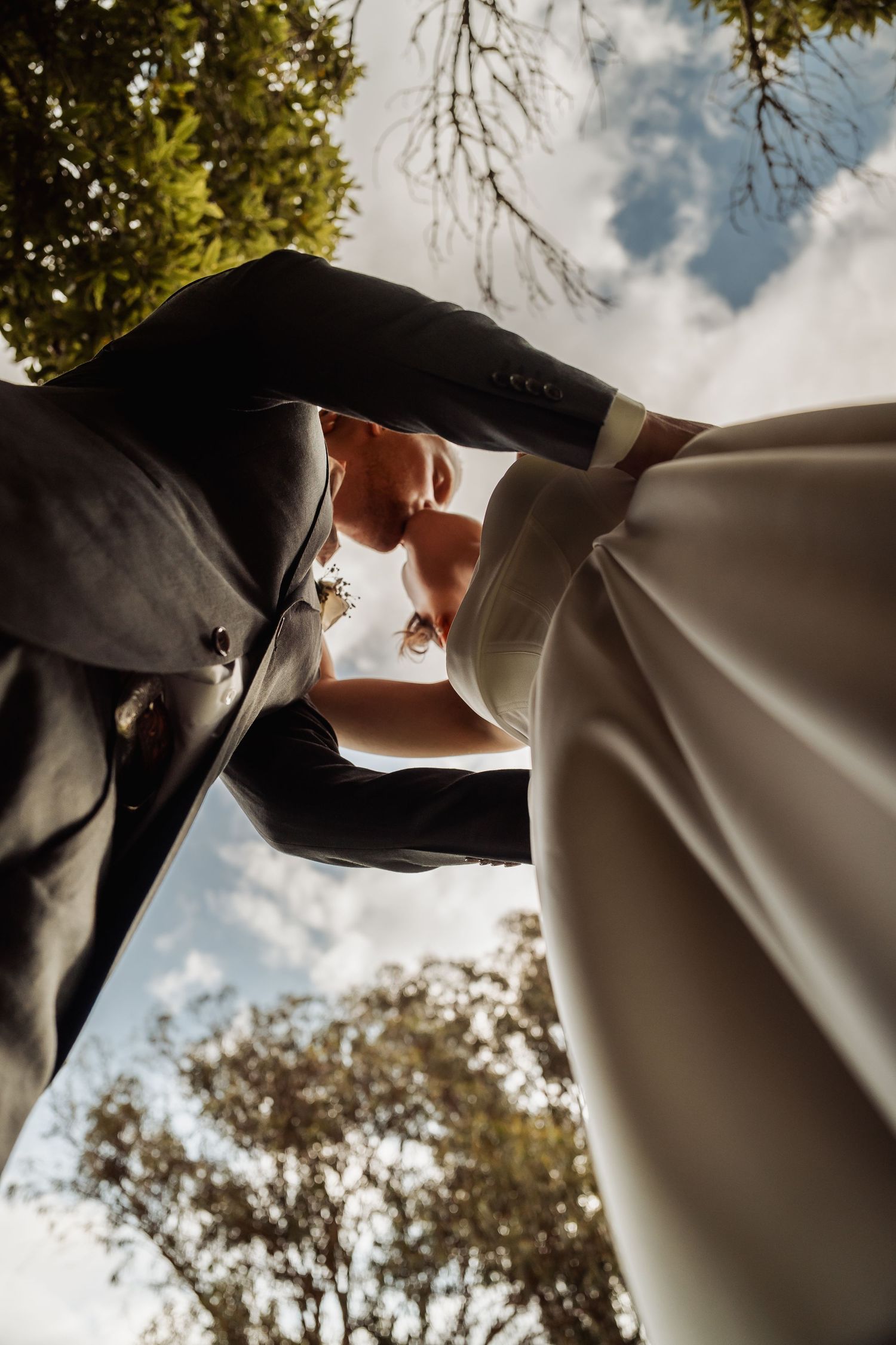 Low-angle artistic shot of bride and groom kissing beneath a canopy of trees against a blue sky.