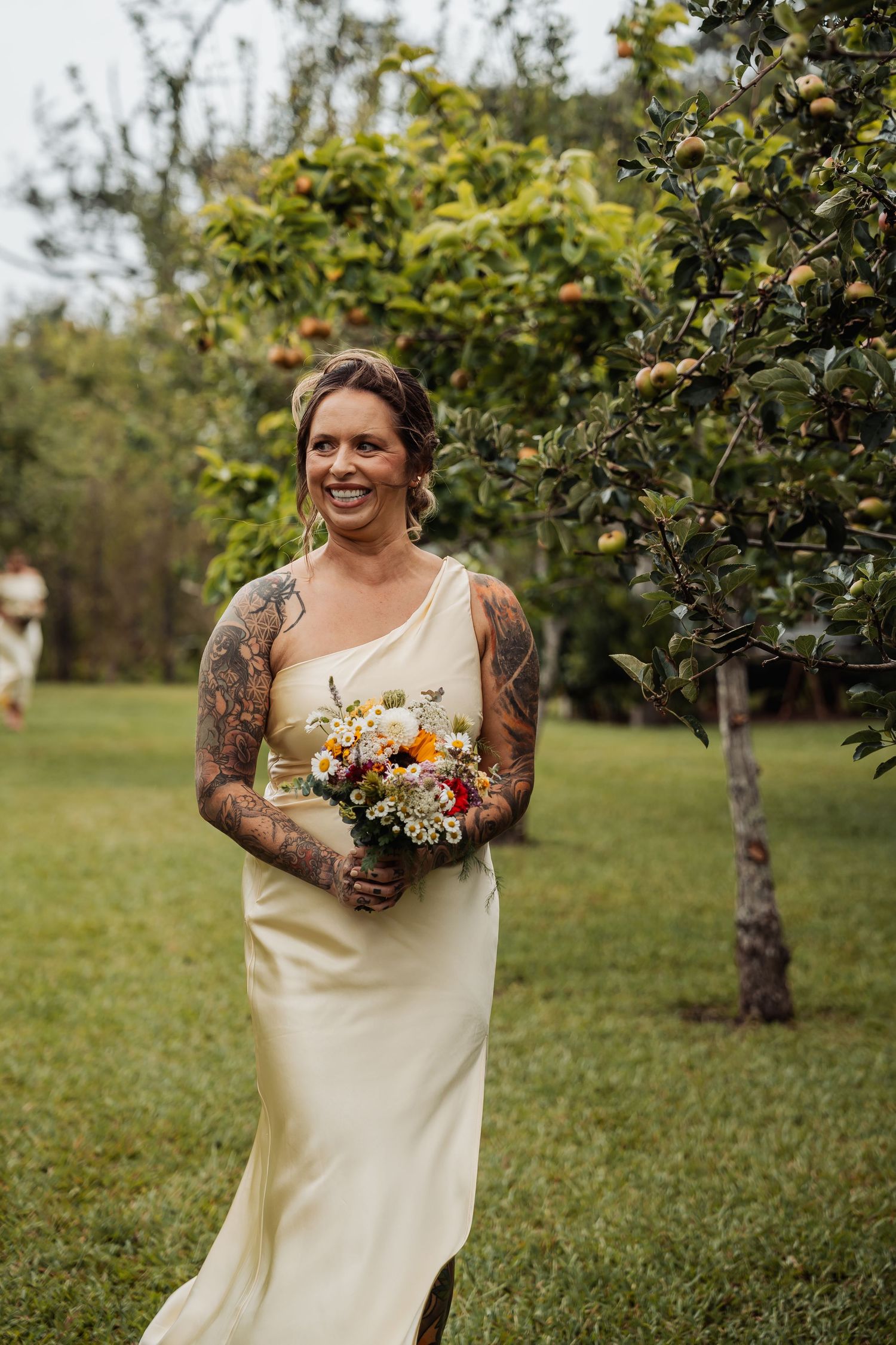 Tattooed bride in ivory gown holding colorful bouquet smiling outdoors near apple tree on wedding day.