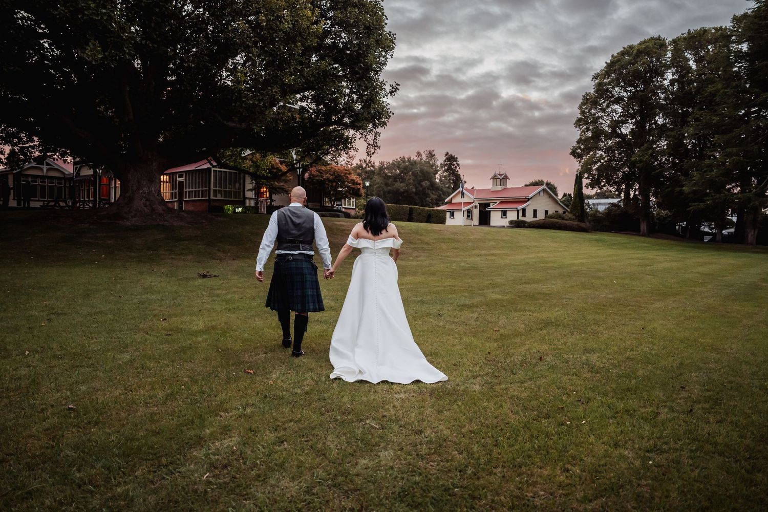 Bride and groom holding hands walking across lawn at sunset in wedding attire and kilt.