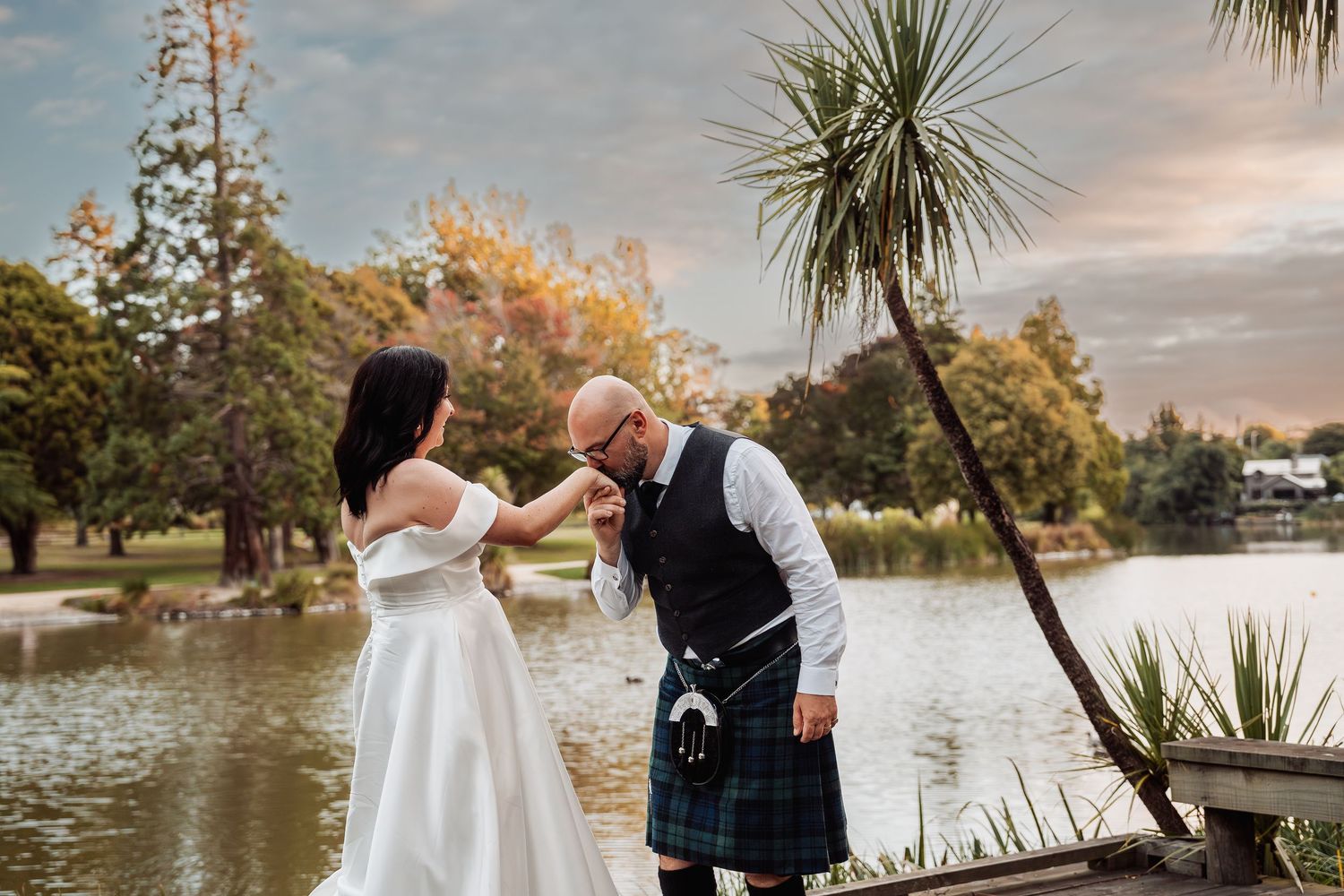 A groom in a tartan kilt kisses his brides hand near a scenic lake at golden hour.