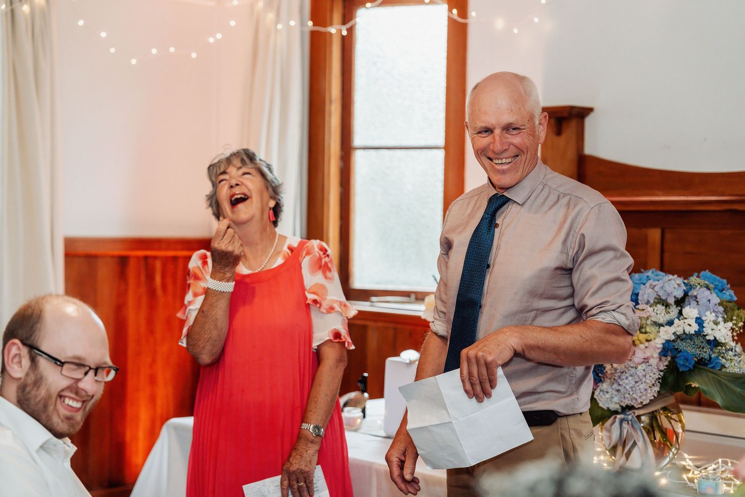 A joyful older couple laughs and smiles during a wedding reception speech inside a decorated venue.