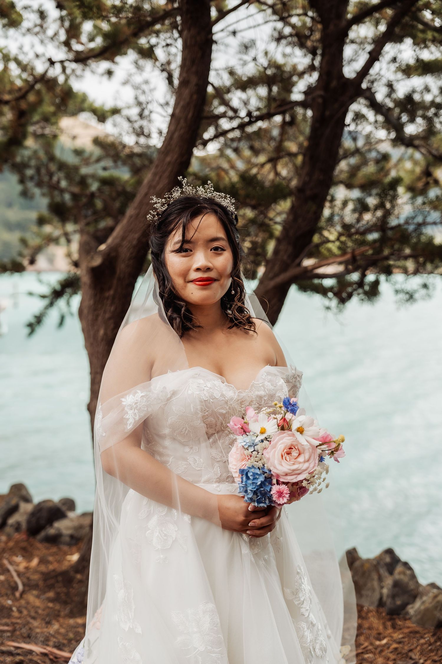 An Asian bride in a white lace off-shoulder gown holds a colorful bouquet near a scenic waterfront backdrop.