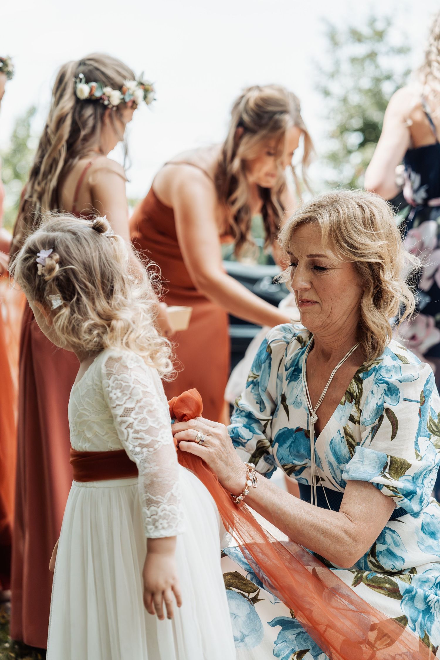 Woman in floral dress adjusts orange sash on flower girl in white lace dress at outdoor wedding.