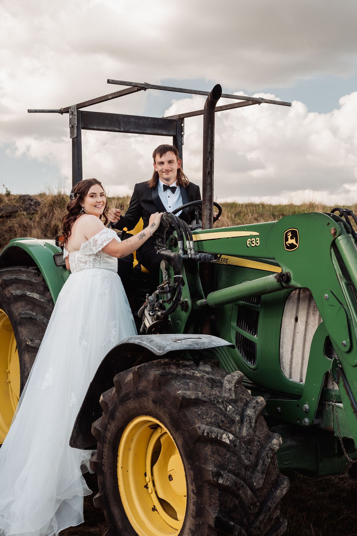 Smiling bride and groom pose together on a green John Deere tractor in an open field.