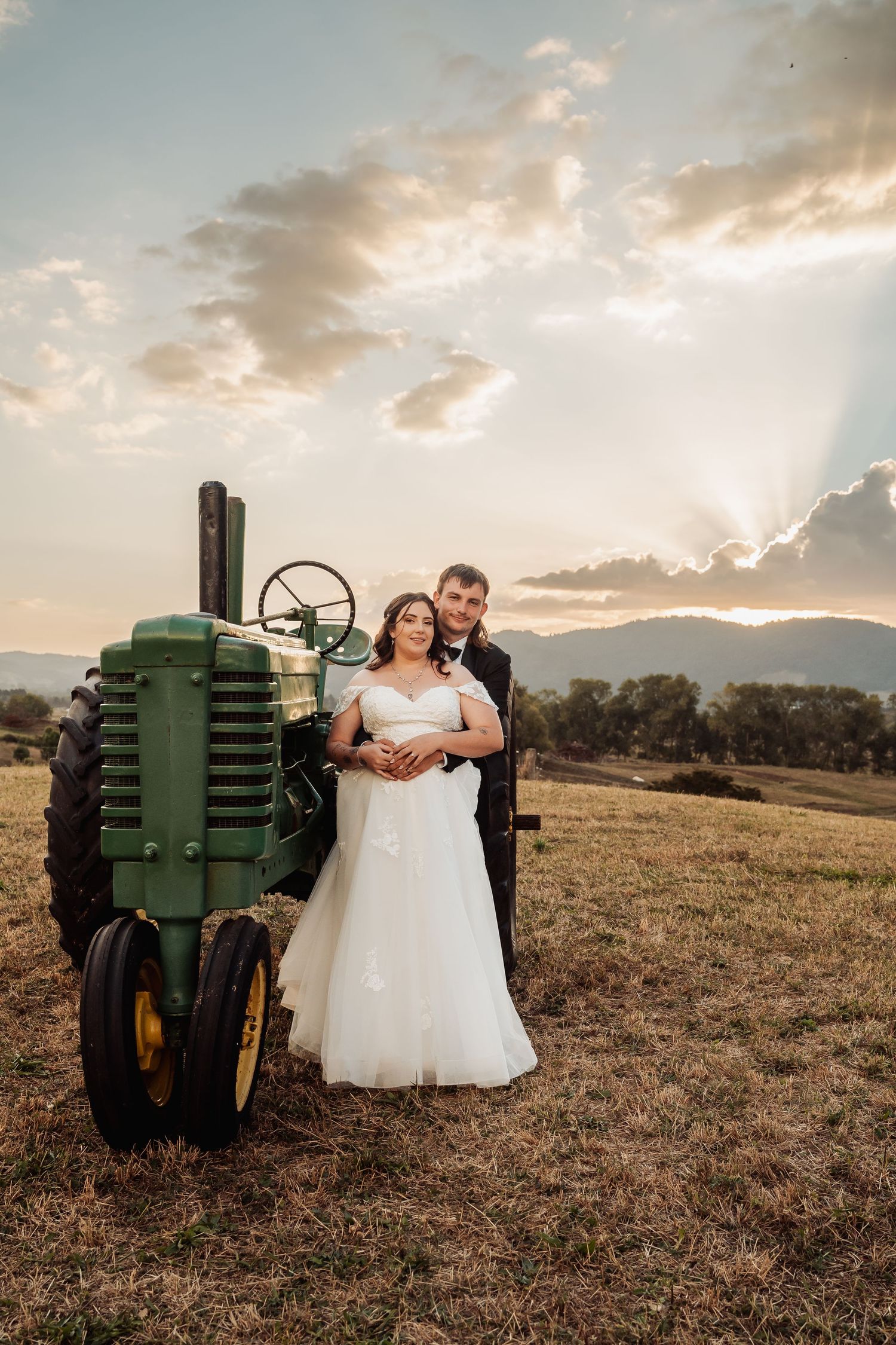 Bride and groom posing beside vintage green John Deere tractor at sunset on rural farm.